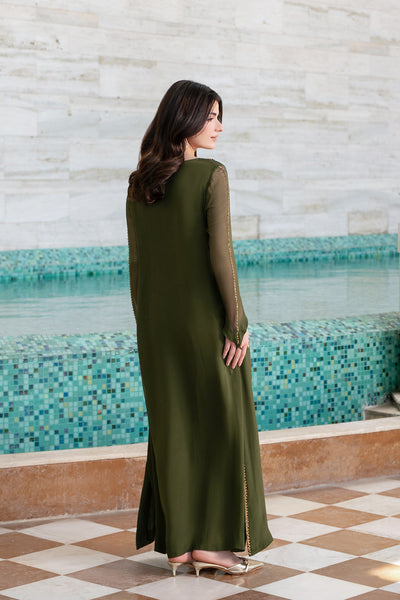 A woman stands with her back to the camera wearing a long olive green dress with sheer long sleeves, standing in front of a tiled pool and a light-colored stone wall wearing a viral dress.